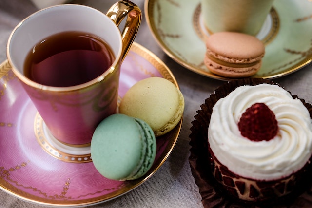 A close up of a pink teacup on a matching saucer next to a cupcake with white icing and a matching green teacup and saucer multicolour macarons are scatrered on the saucers