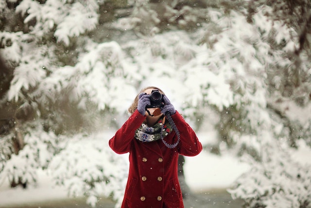 a photographer in a red coat stands in front of snow covered fir trees and takes a photo of falling snow