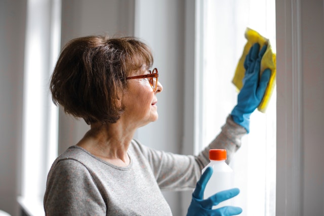 a renter with rusty orange glasses and a short brown bob haircut wears blue latex gloves while cleaning a window with a yellow cloth