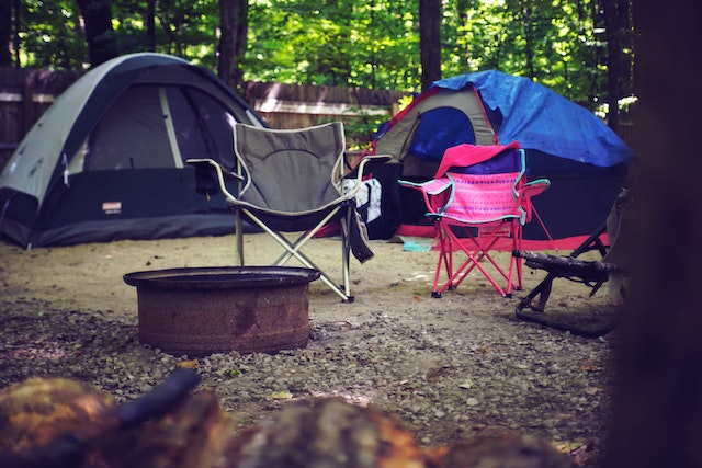 a wooded campsite with two small tents in the background and two camping chairs near a metal fire pit