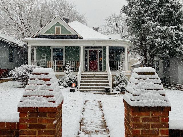snow falls and collects on a green historic bungalow with a wraparound porch