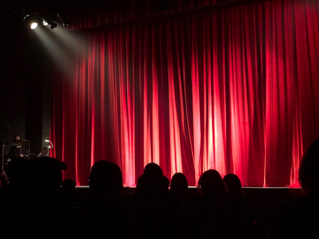 taken from the perspective of the audience various silhouettes of audience members wait for the red curtain to open on stage